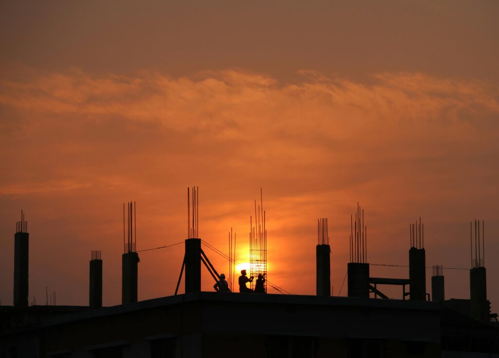Two silhouetted construction workers on a rooftop against a vivid orange and pink sunset, with the sun low on the horizon. The frame of a building is visible with rebar sticking up into the sky.